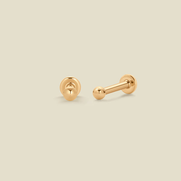 A pair of two gold bead stud earrings, with a spherical ball on one end and a flat back on the other, set against a solid beige background.