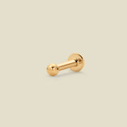 A single gold-colored bead stud earring with a polished spherical ball on one end and a flat back on the other, shown on a solid light beige background.