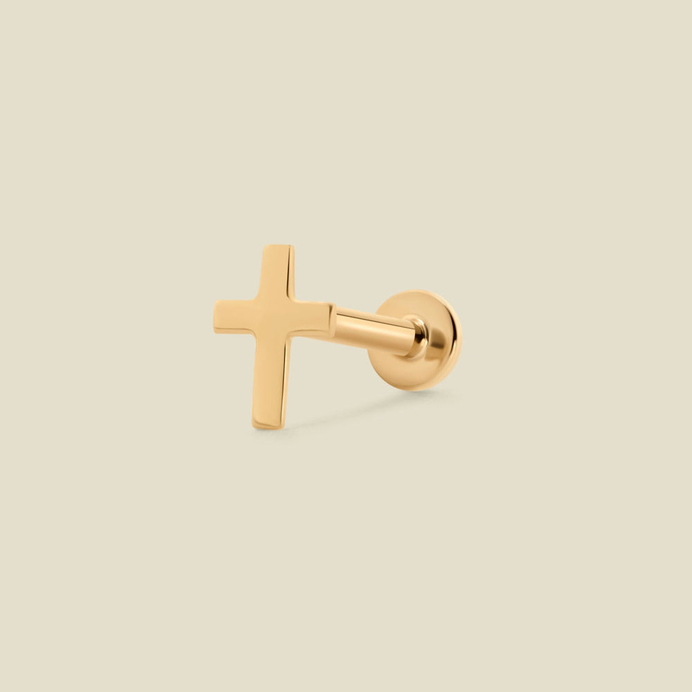 A single gold cross stud earring shown from the side against a solid, light beige background.