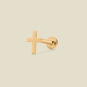A single gold cross stud earring shown from the side against a solid, light beige background.