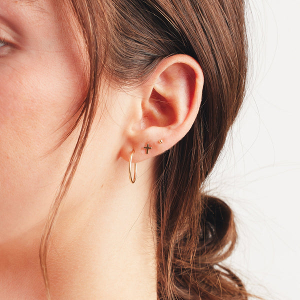 A close up shot of an ear wearing a large thin gold hoop, a gold cross stud, and a gold spherical ball stud.