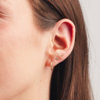 A close up shot of an ear wearing a large thin gold hoop, a gold heart stud, and a gold spherical ball stud.