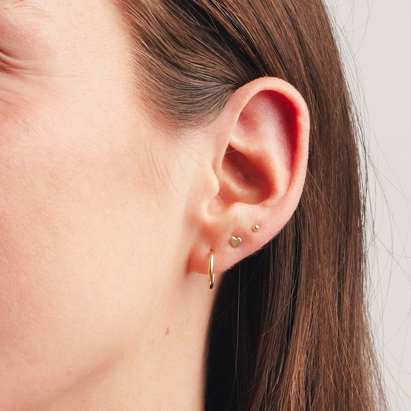 A close up shot of an ear wearing a large thin gold hoop, a gold heart stud, and a gold spherical ball stud.