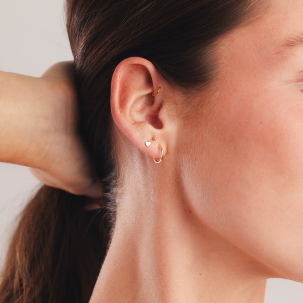 A close up shot of a person's ear with two small thin gold hoops, a gold heart stud, and a gold spherical bead stud.