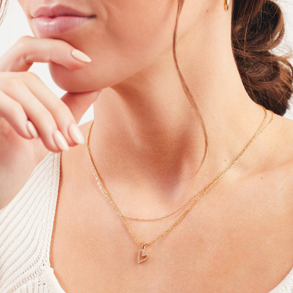 A close up of a person's chest wearing two layered gold necklaces, one with a paperclip link, and the other a pendant necklace with a heart charm featuring small clear stones around the outline.