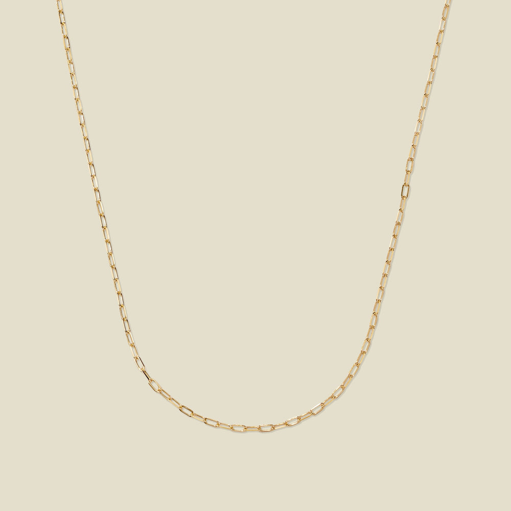 A delicate gold paperclip link chain, set against a solid beige background.