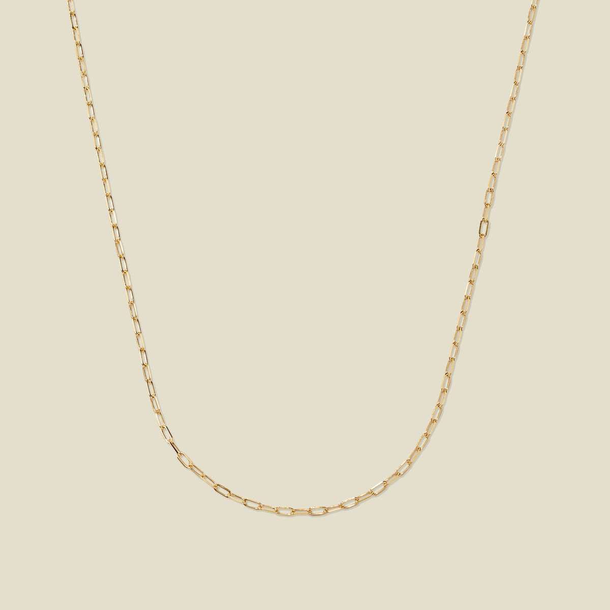A delicate gold paperclip link chain, set against a solid beige background.