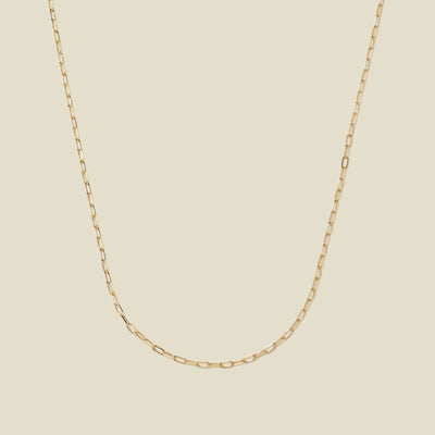 A delicate gold paperclip link chain, set against a solid beige background.
