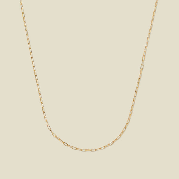 A delicate gold paperclip link chain, set against a solid beige background.
