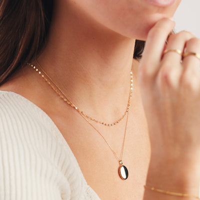 A close up of a person's chest wearing a dainty, textured gold necklace paired with a longer delicate pendant necklace with an oval shaped locket. The person is wearing a white ribbed top, and other gold jewelry on the hand and wrist.