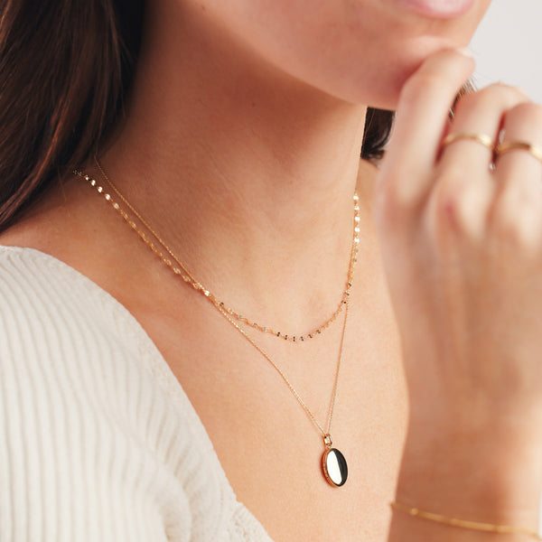 A close up of a person's chest wearing a dainty, textured gold necklace paired with a longer delicate pendant necklace with an oval shaped locket. The person is wearing a white ribbed top, and other gold jewelry on the hand and wrist.