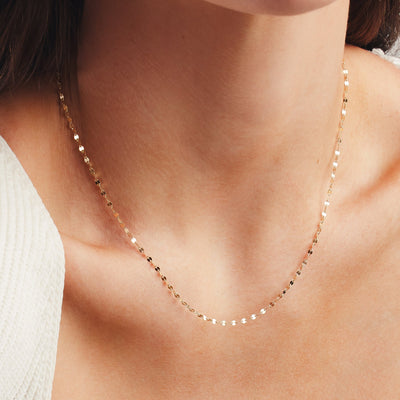 A close up shot of a person's neck wearing a delicate, textured gold chain necklace and a white knit top.