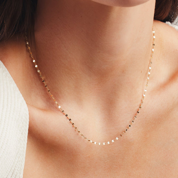 A close up shot of a person's neck wearing a delicate, textured gold chain necklace and a white knit top.