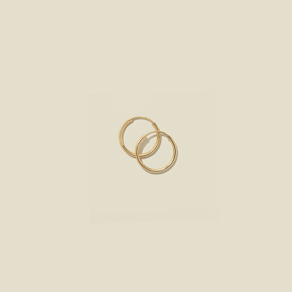 A pair of thin gold seamless hoop earrings, stacked on one another, set against a solid beige background.