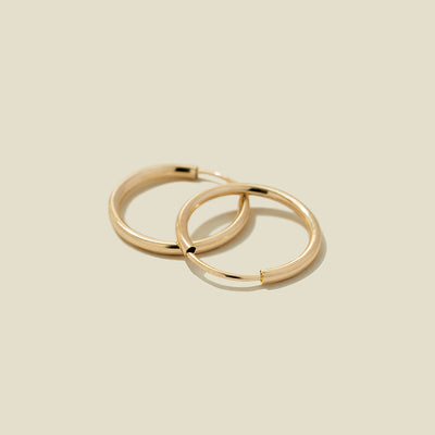 A pair of thin gold seamless hoop earrings, stacked on one another, set against a solid beige background.