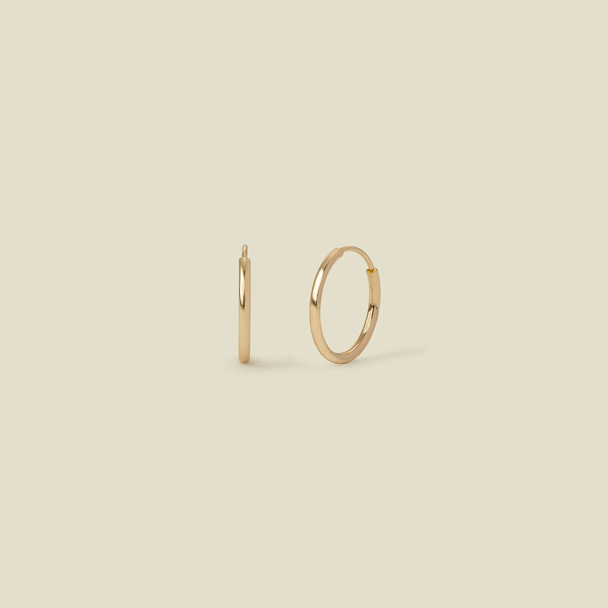 A pair of thin gold seamless hoop earrings, standing vertically, set against a solid beige background.