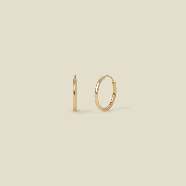 A pair of thin gold seamless hoop earrings, standing vertically, set against a solid beige background.