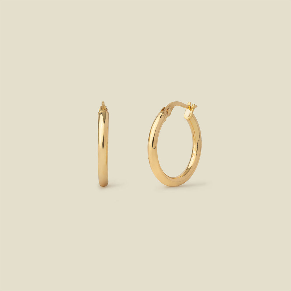A pair of polished gold hoop earrings shown on a plain warm beige background, with one earring facing forward and the other in side view.
