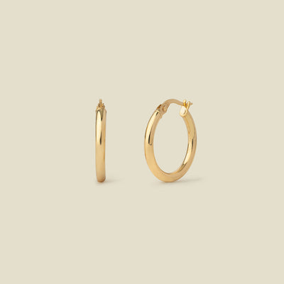 A pair of polished gold hoop earrings shown on a plain warm beige background, with one earring facing forward and the other in side view.
