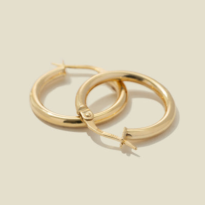 A pair of gold polished hoop earrings, stacked on top of one another, set against a warm, solid beige background.