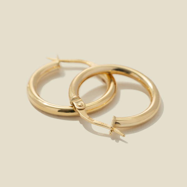 A pair of gold polished hoop earrings, stacked on top of one another, set against a warm, solid beige background.