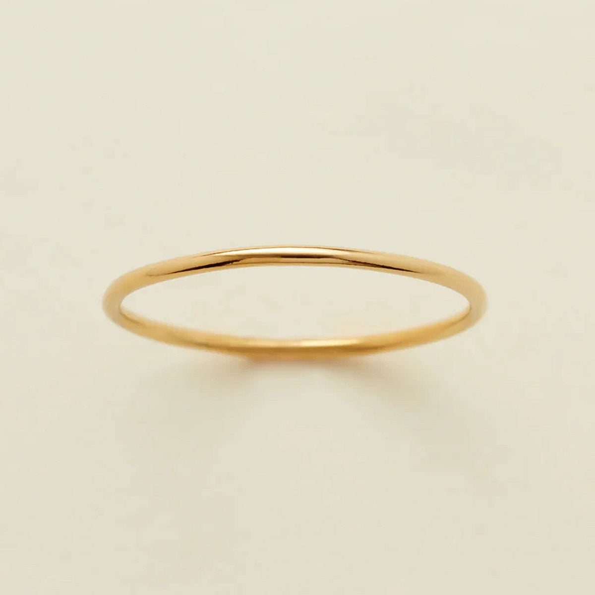 A minimalist, thin yellow gold band ring with a polished finish, displayed on a clean, solid off-white background.