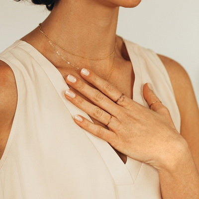 A lifestyle photo featuring a person's chest with hand laying across it, wearing multiple gold wire rings, and layered gold necklaces, along with a cream colored v-neck blouse.