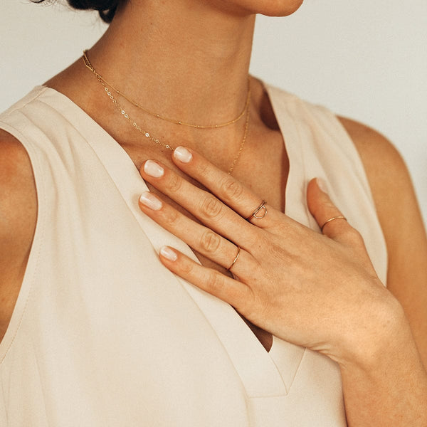 A lifestyle photo featuring a person's chest with hand laying across it, wearing multiple gold wire rings, and layered gold necklaces, along with a cream colored v-neck blouse.