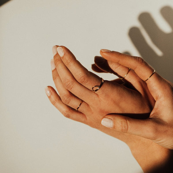 A close up photo of two hands wearing multiple gold wire rings, set against a warm, cream background with shadowing.