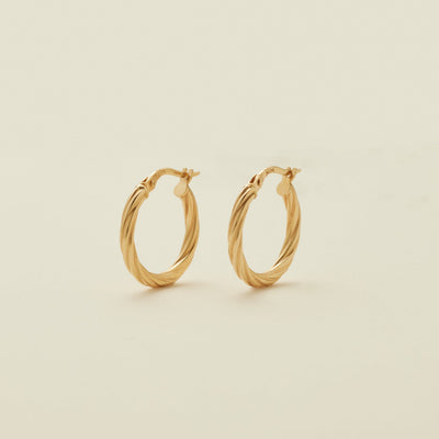 A pair of two polished gold hoops with a twist, textured design, standing upright against a solid beige background.