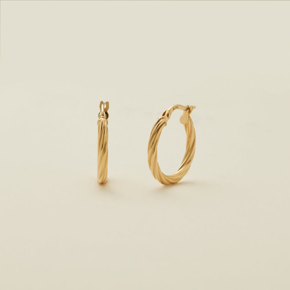 A pair of two polished gold hoops with a twist, textured design, standing upright against a solid beige background.