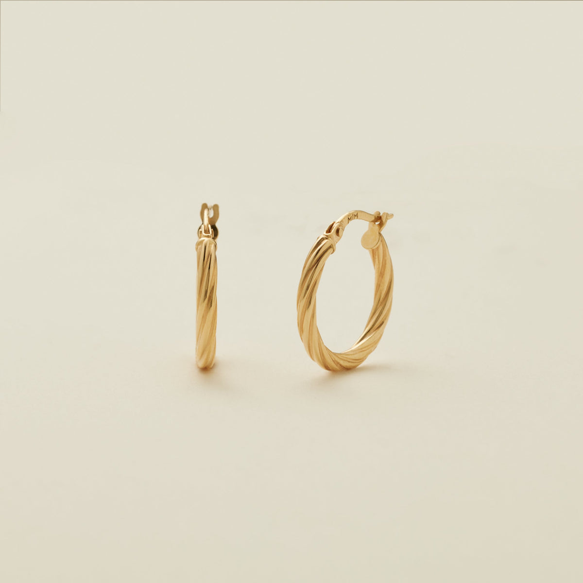 A pair of two polished gold hoops with a twist, textured design, standing upright against a solid beige background.
