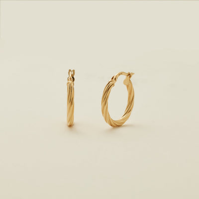 A pair of two polished gold hoops with a twist, textured design, standing upright against a solid beige background.