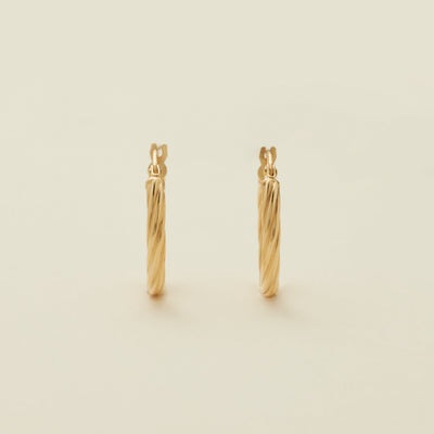 A pair of two polished gold hoops with a twist, textured design, standing upright against a solid beige background.