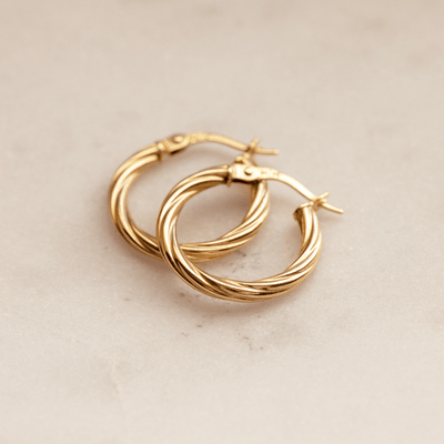 A pair of two polished gold hoops with a twist, textured design, laying on top of each other, against a textured beige background.