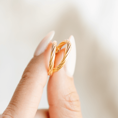 A person holding a pair of two polished gold hoops with a twist, textured design between two fingers, set against a clean, light background.