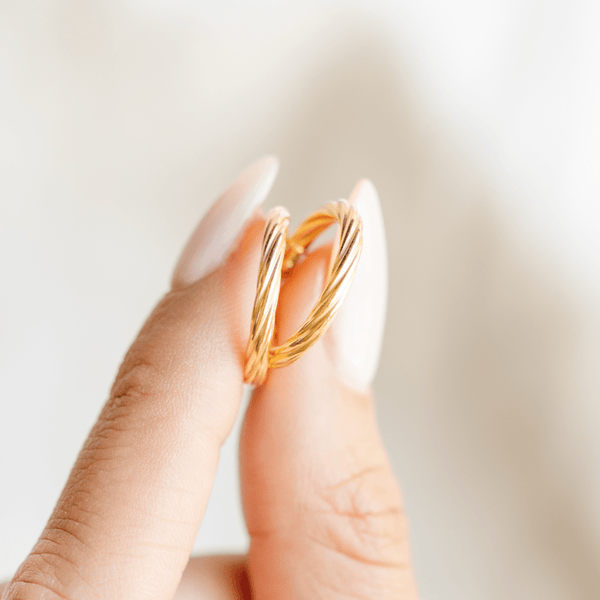 A person holding a pair of two polished gold hoops with a twist, textured design between two fingers, set against a clean, light background.