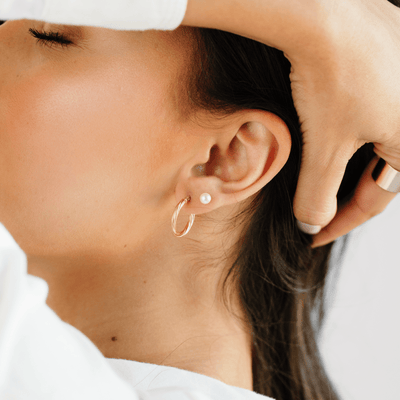 A close up lifestyle image of a person's profile, wearing a gold, twist textured hoop earring and a pearl stud.