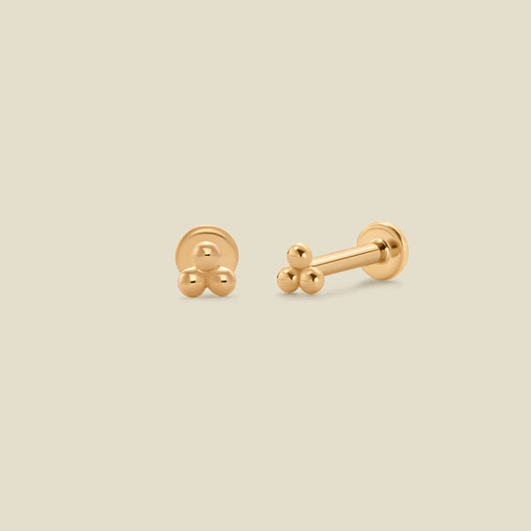 A pair of yellow-gold stud earrings, with a trio small, round beads in a triangular grouping, and a flat backing, set against a solid beige background.