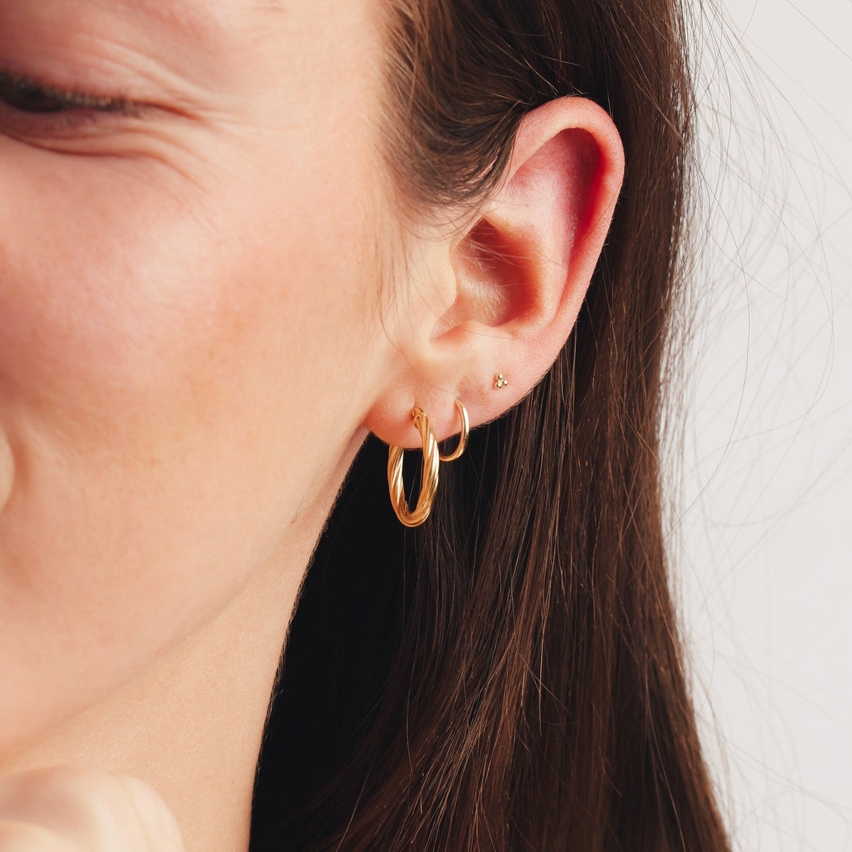 A lifestyle photo of a person's ear wearing a large, gold twisting hoop, a small, thin gold hoop, and a gold stud with three spherical beads.