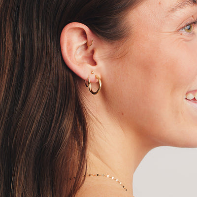 A lifestyle photo of a person's ear wearing a large, gold twisting hoop, two small, thin gold hoop, and a gold stud with three spherical beads.