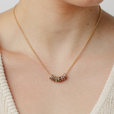 A close up lifestyle photo of a person wearing a gold pendant chain necklace with round, gold charms featuring square bezel-set stones side-by-side, each charm with a different color stone, set against a solid beige background.