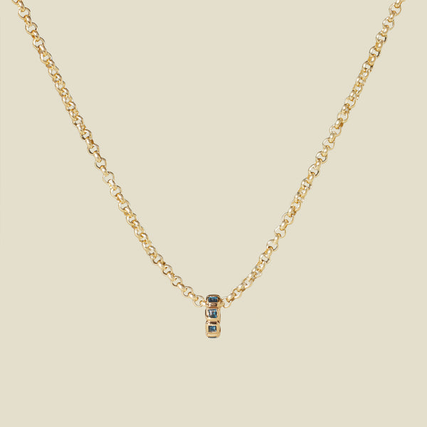 A gold pendant chain necklace with a round, gold charm featuring square bezel-set blue-green stones side-by-side, set against a solid beige background.