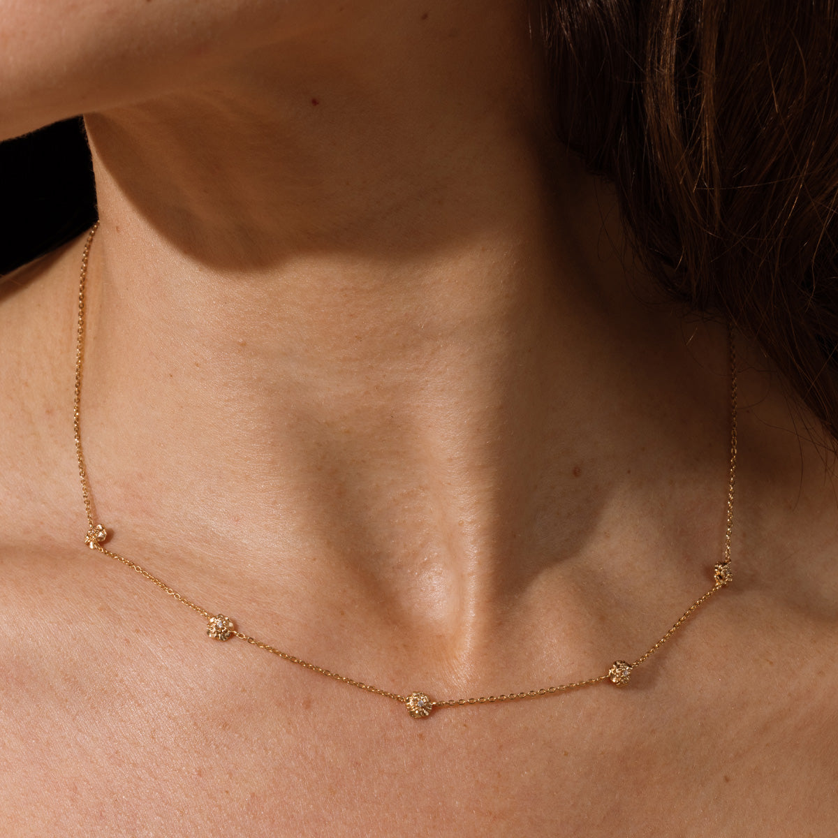 A lifestyle image of a dainty gold necklace on a person's neck with stationary textured carnation flowers with small accent stones in the center along the chain.