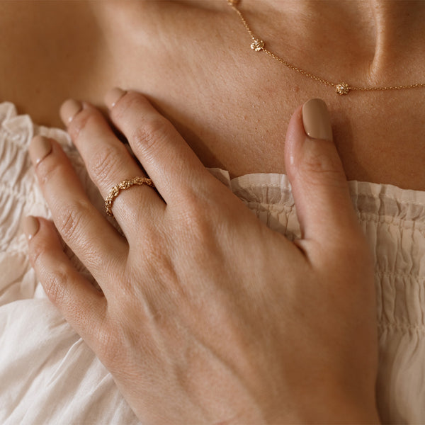 Close up of a hand with a gold floral-etched ring band with textured carnation flowers and small clear gemstones, with the matching necklace being worn in the background.