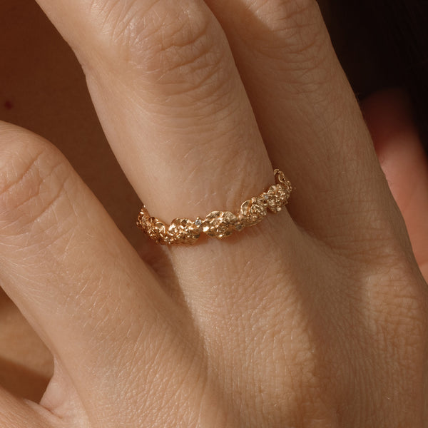 Close up of a hand with a gold floral-etched ring band with textured carnation flowers and small clear gemstones.