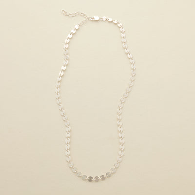 A delicate silver necklace with flat, polished circular discs linked together, set against a solid beige background.