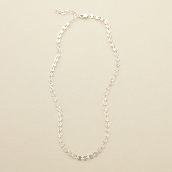 A delicate silver necklace with flat, polished circular discs linked together, set against a solid beige background.