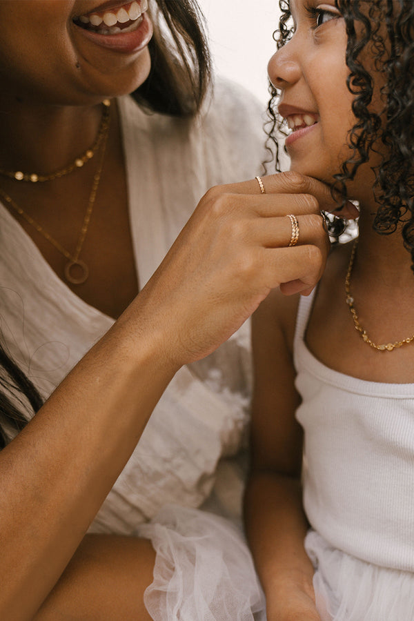 A close up lifestyle photo of an adult woman tenderly putting her hand under the chin of a young girl, with both of them wearing dainty gold jewelry and white outfits.