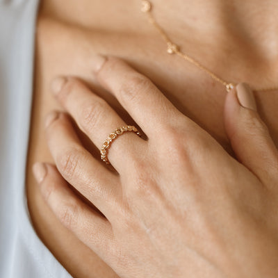 Close up of a hand with a gold floral-etched ring band with textured violet flowers and small clear gemstones, with a matching necklace being worn in the background.